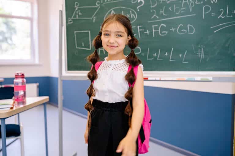 Young girl with a pink backpack standing in front of a chalkboard at school.